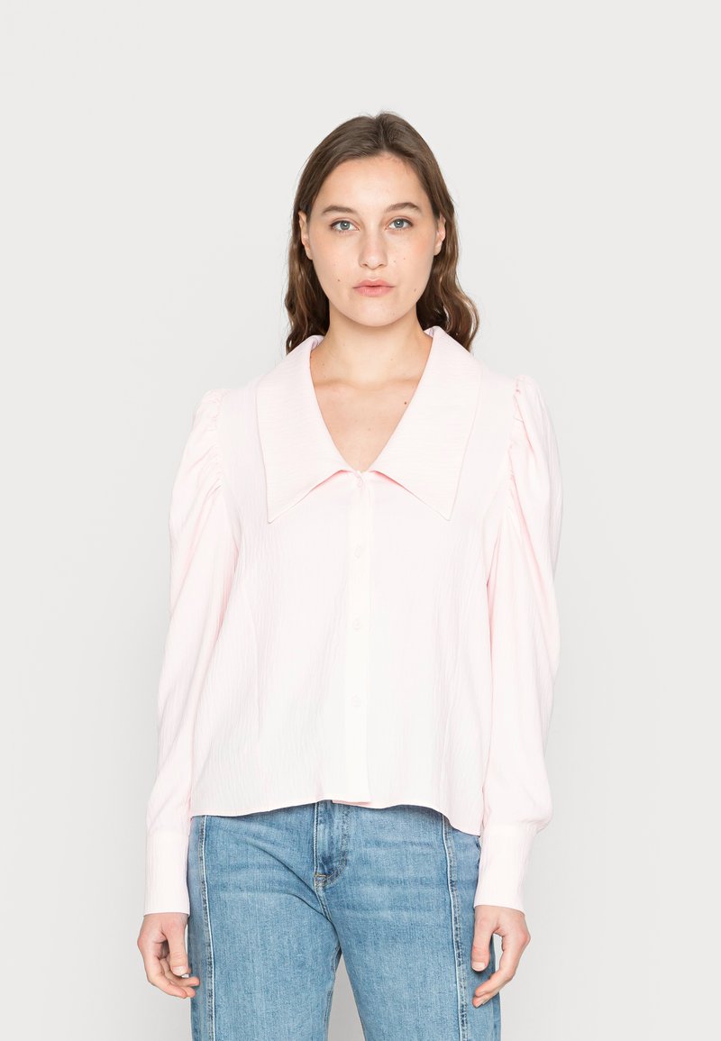 Camisa de botão rosa claro com um grande colarinho, mangas bufantes e tecido texturizado. Combinada com jeans azul claro.