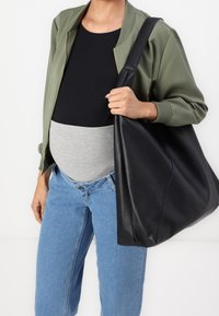 Osoba v modrých džínách, černém topu a olivovém kabátu, nese velkou černou texturovanou kabelku přes rameno.