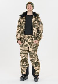 Camouflage-patroon ski-jack en broek in beige en groen, gemaakt van waterdicht materiaal, gecombineerd met een donker langemouwenshirt en zwarte skischoenen.
