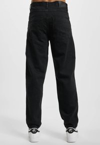 Schwarze Jeans aus Denim mit geradem Bein, ausgestattet mit zwei Gesäßtaschen und einem markierten Taillenbund. Dazu werden weiße Sneakers getragen.