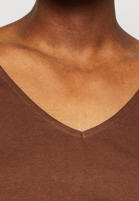 Top marrón de cuello en V hecho de una tela suave. Costuras visibles alrededor del escote. El tono y la textura de la piel en el área del cuello también son visibles.