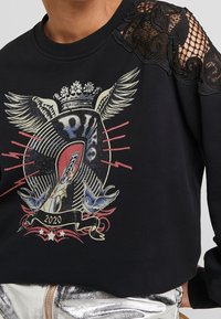 Felpa nera con dettaglio in pizzo sulle spalle, stampa grafica di una corona alata, uccelli e un banner con "2020", indossata con pantaloni argentati metallici.