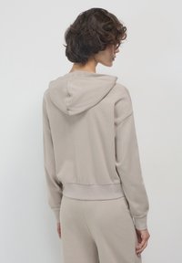 Donna con capelli ricci corti, che indossa un hoodie beige e pantaloni coordinati, girata di lato contro uno sfondo chiaro e neutro.