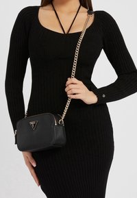 Borsa nera con texture e tracolla in catena dorata, dotata di un logo triangolare. La borsa ha una forma rettangolare ed è chiusa da una zip.