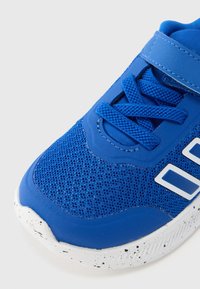 Pantof sport albastru cu partea superioară din plasă, capăt de deget din cauciuc și talpă exterioară albă cu pete. Are șireturi albastre și o curea cu velcro pentru o fixare sigură.