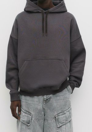 Dunkelgrauer oversized Hoodie mit vorderer Kängurutasche und Kordelzügen, getragen zu hellgrauen Cargo-Jeans mit großen aufgesetzten Taschen.
