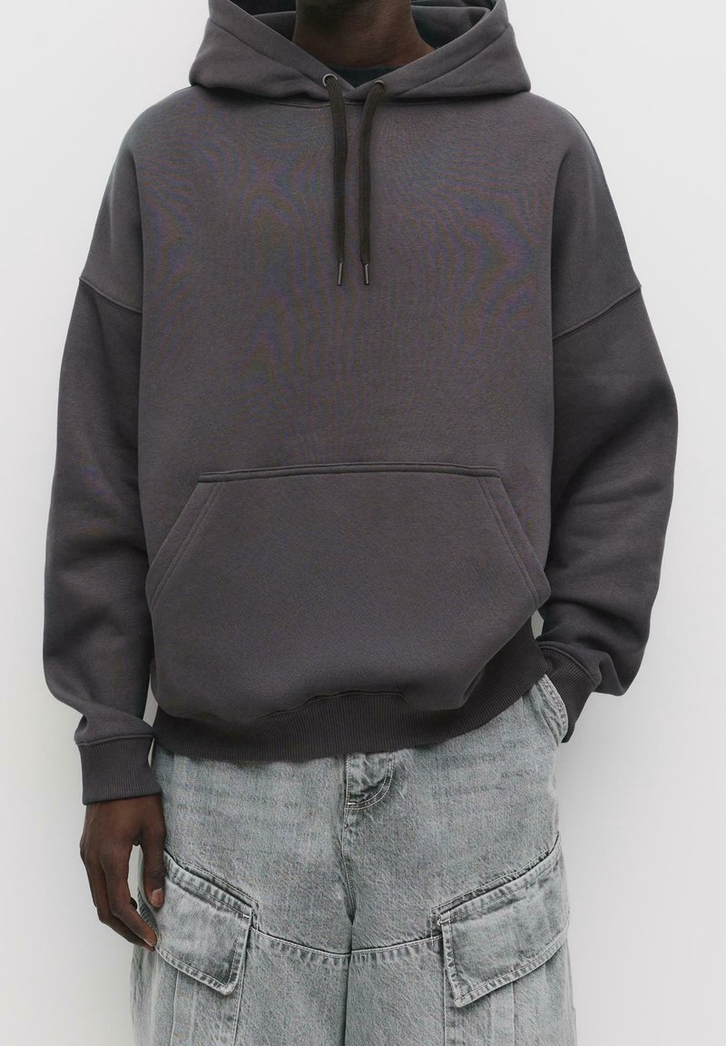 Sweat à capuche oversize gris foncé avec poche kangourou à l'avant et cordons, porté avec un jean cargo gris clair avec de grandes poches plaquées.