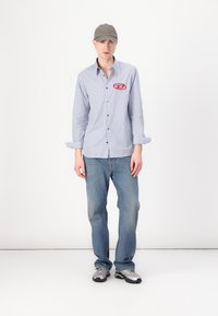 Striped long-sleeve shirt in blue and white with button closure and red logo, paired with light blue denim jeans and grey athletic shoes.