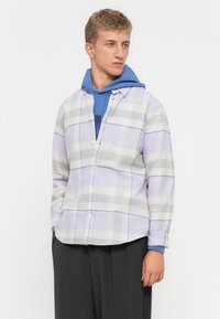 Joven que lleva una sudadera azul debajo de una camisa de cuadros en tonos púrpura pastel y gris, combinada con pantalones de pierna ancha gris oscuro, mirando hacia la derecha.