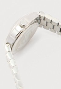 Ασημένια μεταλλική κάσα ρολογιού με σκαλιστό πίσω μέρος και αλυσίδα από μέταλλο, δείχνοντας τη πλευρική όψη που περιλαμβάνει το κορώνα και μέρος του κλιπ της αλυσίδας.