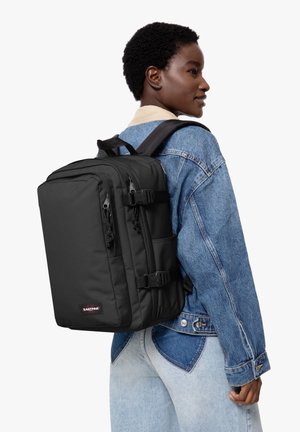 Mochila Eastpak negra con múltiples compartimentos, correas ajustables y cremalleras, llevada en la espalda de una persona con una chaqueta de mezclilla.