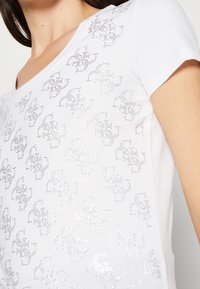 Camiseta blanca de cuello en V con mangas cortas, decorada con un patrón repetitivo de pequeños diseños brillantes de pedrería en el pecho y el hombro.