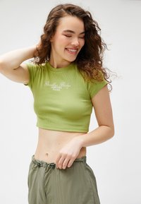 Top corto verde con canalé, de mangas cortas y cuello redondo, que presenta el texto "Brooklyn Bridge New York" en blanco. Combinado con pantalones holgados olivo.