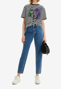 Musta-valkoraidallinen T-paita, jossa värikäs graafinen kuvio, yhdistetty korkeavyötäröisiin sinisiin farkkuihin ja valkoisiin lenkkareihin, kädessä musta laukku.