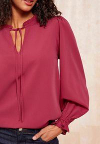 Blusa bordeaux con scollatura a oblò, colletto arricciato e polsini elasticizzati. Realizzata in tessuto morbido, presenta una vestibilità comoda e maniche fluide.