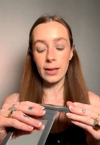 Jeune femme aux longs cheveux bruns tenant et ajustant un miroir compact de maquillage sur un fond uni.