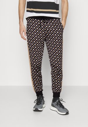 Mann trägt schwarz-braune gemusterte Jogginghosen mit Kordelzug in der Taille, schwarzen Bündchen und grauen Sneakers und steht vor einem weißen Hintergrund.