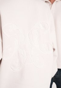 Lyserød sweatshirt med et tekstureret, hvidt broderet blomsterdesign på ryggen. Blødt, glat stof med en afslappet pasform.