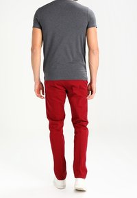 Hombre de pie, vistiendo una camiseta gris ajustada de manga corta, pantalones rojos y zapatillas blancas, de espaldas en un fondo blanco liso.