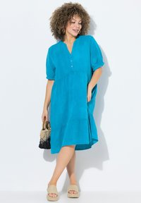Teal Kurzarmkleid mit V-Ausschnitt, Knöpfen, gestuftem Design und lockerer Passform. Kombiniert mit beigen Flip-Flops und einer geflochtenen Tasche.