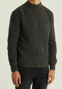 Gestrickter dunkelgrüner Pullover mit hohem Kragen, strukturiertem Muster, tailliertem Design und einem kleinen schwarzen Markenlogo auf der Brust.