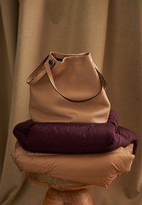 Beige Leder-Bucket-Bag mit glatter Textur und schwarzem Riemen, platziert auf zwei gepolsterten Lagen – eine bordeauxfarben und eine beige – vor neutralem Hintergrund.