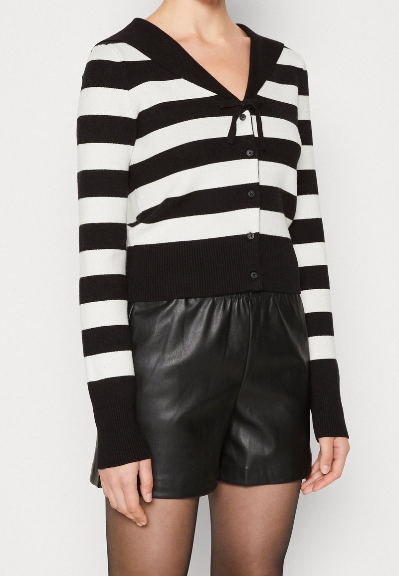 Cardigan à rayures noires et blanches avec encolure en V et un détail de nœud au col, doté d'une fermeture à boutons et de poignets côtelés. Associé à un short en cuir.