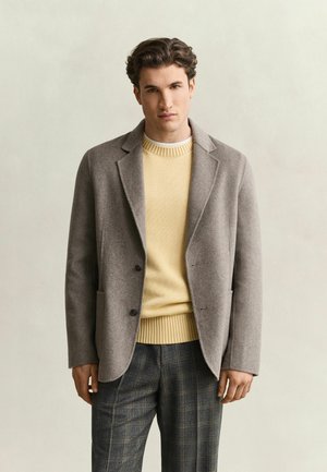 Jonge man met kort, golvend haar die een beige jas over een gele trui en grijze geruite broek draagt, staand tegen een effen achtergrond.