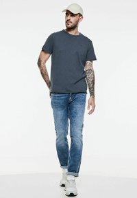 Tmavě šedé tričko s kulatým výstřihem v kombinaci s vybledlými modrými džíny a bílými teniskami. Model má na sobě světlý béžový klobouk. Tetování viditelné na pažích.