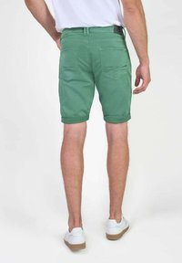 Groene shorts met een opgerolde zoom, achterzakken en een label op de tailleband. Draagt witte sneakers op een effen achtergrond.