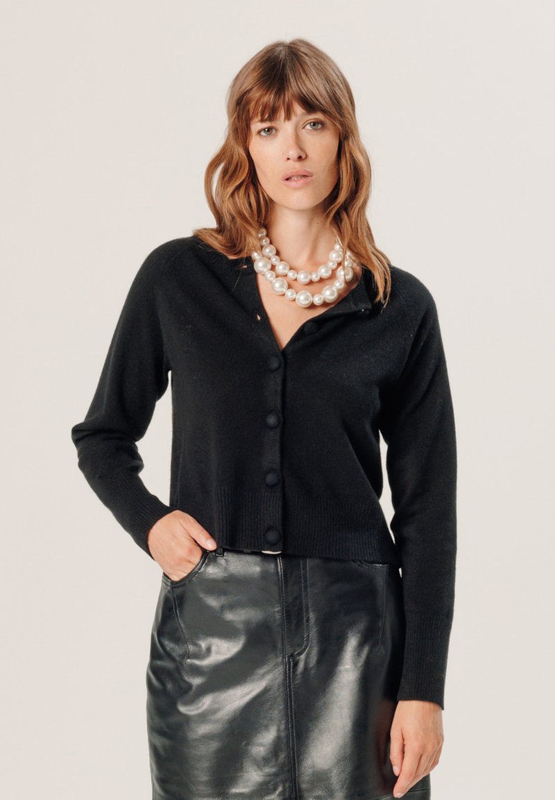 Schwarze Strickjacke mit tailliertem Design, langen Ärmeln, Knopfverschluss und geripptem Saum, kombiniert mit einem schwarzen Lederrock. Perlen als Accessoires.