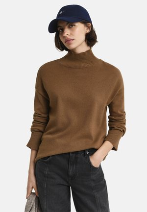 Vrouw in een bruin coltrui, zwarte jeans en een marineblauwe pet, staande met één hand in de zak en de andere hand met stof.