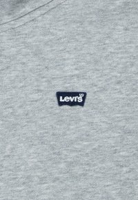 Tela de algodón gris con una textura suave. Presenta un pequeño parche con el logo de Levi's en azul marino en la esquina superior izquierda. Diseño simple y casual.
