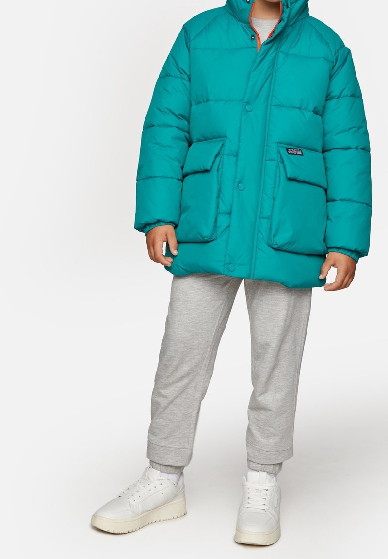 Tyrkysová péřová bunda s kapucí, dvěma velkými předními kapsami a zapínáním na knoflíky. Nošena přes šedé tepláky a bílé tenisky.