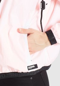 Jasnoróżowa kurtka przeciwdeszczowa z czarnym elastycznym paskiem w talii, zapinanymi na zamek kieszeniami bocznymi i kapturem ze sznurkiem. Posiada teksturowaną tkaninę i metkę z logo.