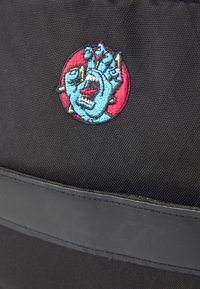 Črn nahrbtnik iz tkanine s pisanimi izvezenimi našitki roke s obrazom, na rdeči krožni podlagi, z črnimi kovinskimi dodatki.