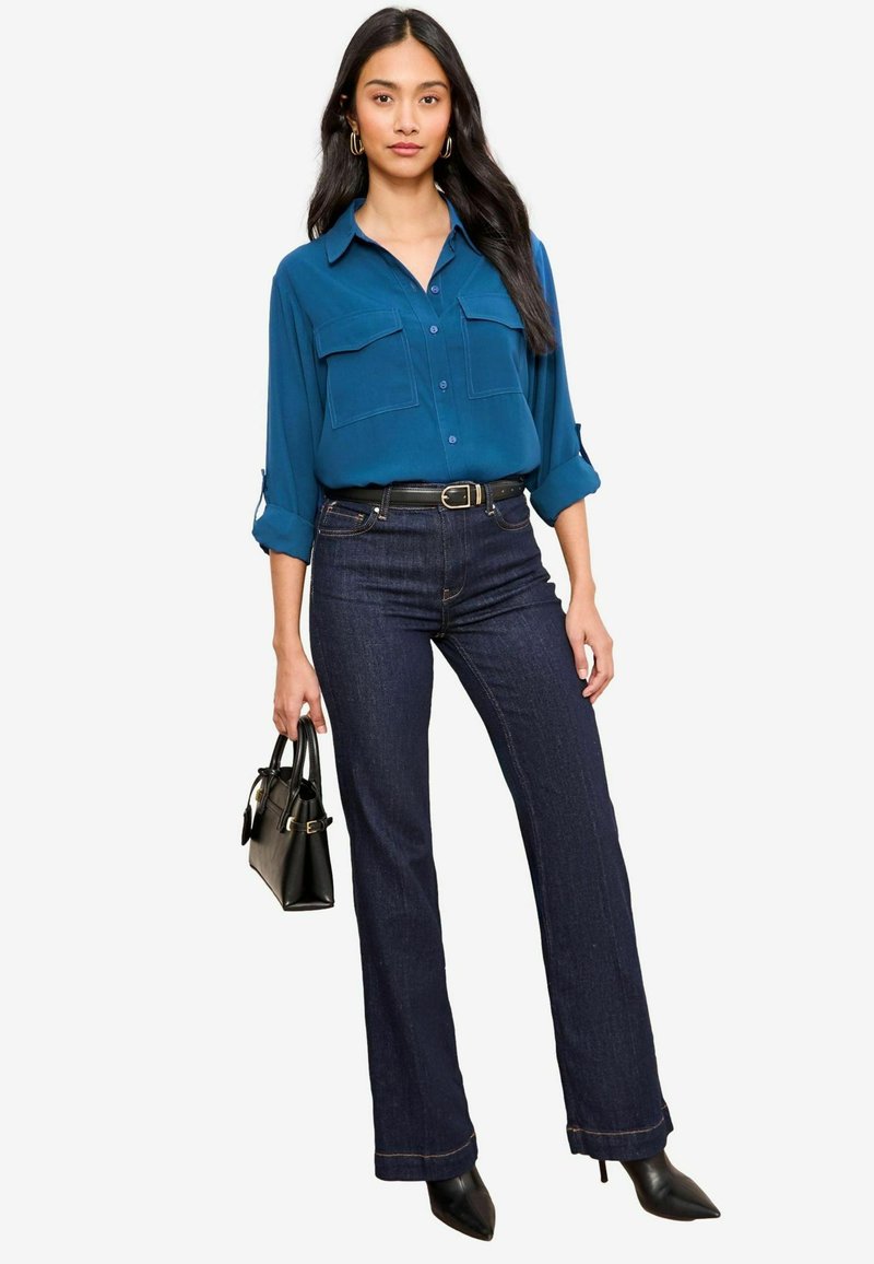 Teal skjorte med knapper og to brystlommer, mørk denim flared jeans, sort bælte, sort håndtaske og spidse hæle.