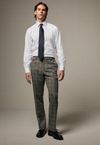 Weißes Hemd, navyblauer Krawatte mit Muster, graue karierte Hose und schwarze Schuhe. Die Hose hat eine vordere Taschendetail.