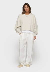 Felpa cropped beige chiaro con collo rotondo e maniche arricciate, indossata sopra una camicia bianca; abbinata a pantaloni bianchi larghi a vita alta.