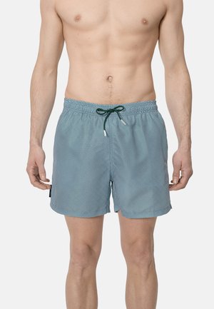 Hombre con pantalones cortos de baño azules con estampado y cordón verde oscuro, de pie contra un fondo claro y liso.