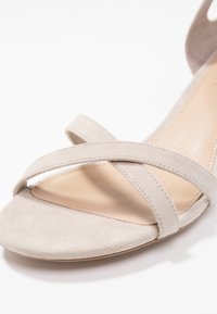 Béžové semišové sandály s otevřenou špičkou, se dvěma tenkými křížovými pásky a hladkou stélkou, vystavené na bílém pozadí.