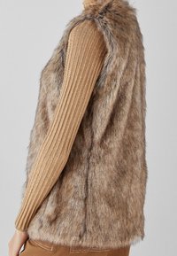 Hnědá vesta z umělé kožešiny s vysokým límcem, měkká na dotek a bez rukávů, doplněná žebrovaným béžovým svetrem s dlouhým rukávem.