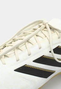 White soccer cleat featuring a textured upper, beige laces, black accents with zigzag patterns, and gold detailing along the sides.