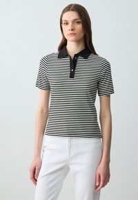 Polo a maniche corte a righe bianche e nere con colletto nero, tre bottoni dorati e una vestibilità aderente. Indossato con pantaloni bianchi.
