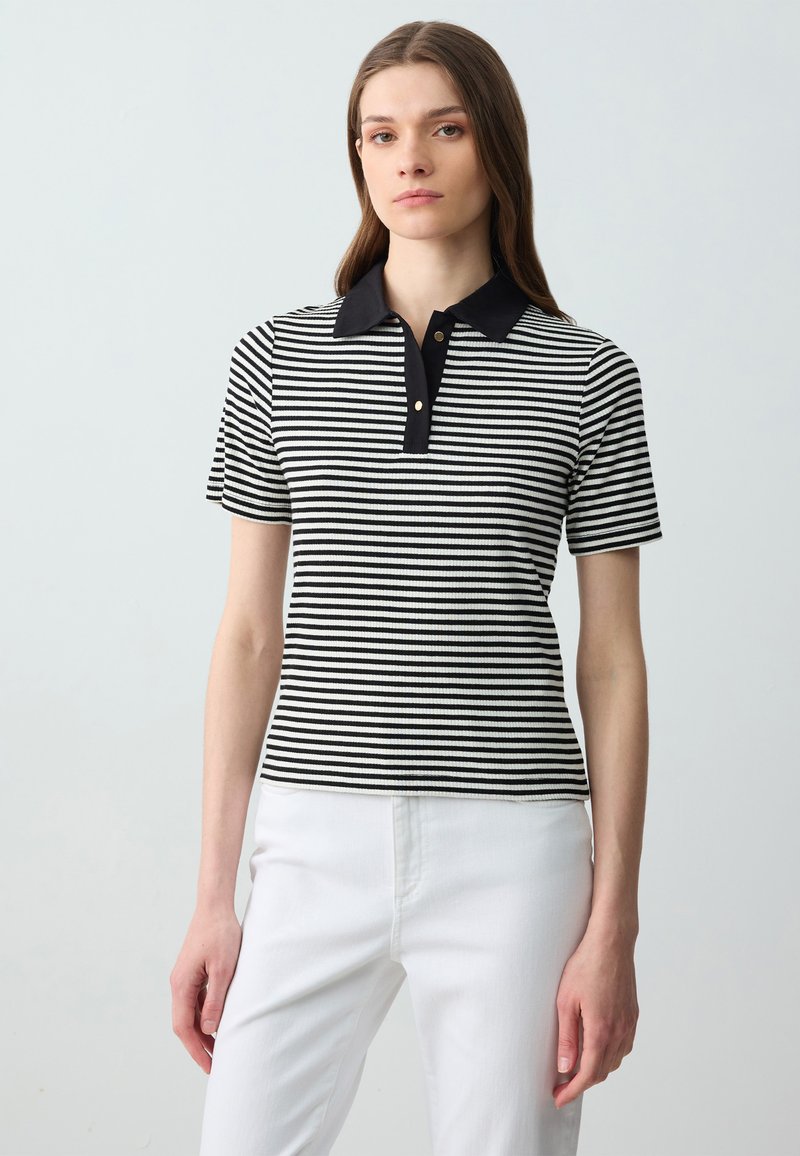 Polo a maniche corte a righe bianche e nere con colletto nero, tre bottoni dorati e una vestibilità aderente. Indossato con pantaloni bianchi.
