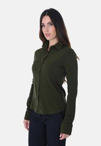Camicia a maniche lunghe button-up verde scuro con colletto classico, texture liscia e vestibilità su misura. Presenta un orlo dritto e chiusura con bottoni.