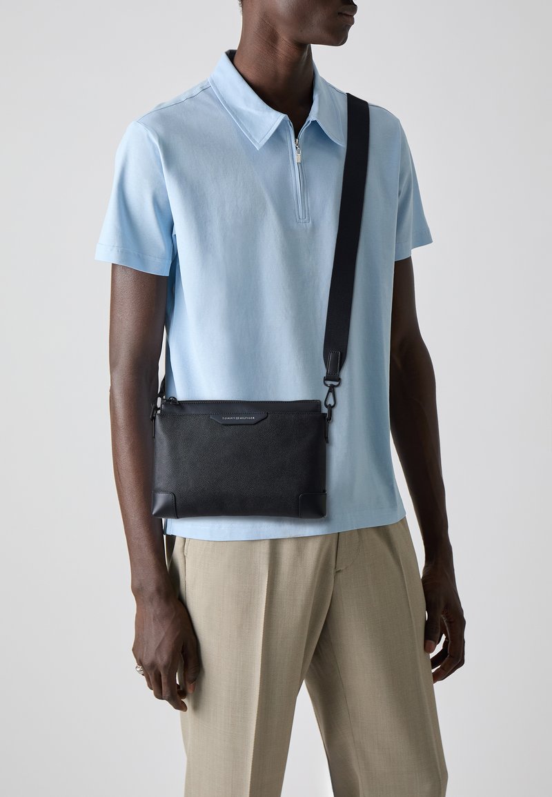 Black leather crossbody bag with a textured finish, designed with a zipper closure and adjustable strap, worn over a light blue polo shirt.