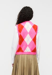 Πλεκτό γιλέκο με πολύχρωμο  σχέδιο αργκόιλ σε ροζ, κόκκινο και άσπρο, με ριμπ ροζ τελείωμα. Φορεμένο πάνω από λευκό πουκάμισο με μακρύ μανίκι.