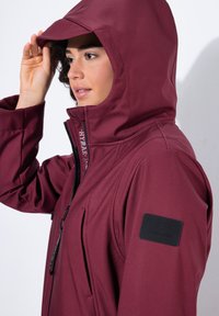 Chaqueta impermeable color burdeos con un cuello alto y capucha ajustable. Presenta una etiqueta negra en el hombro y un cierre de cremallera. Textura suave.