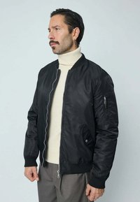 Veste bombardier noire avec fermeture éclair à l'avant, poignets côtelés et une poche sur la manche gauche ; portée sur un col roulé crème. Texture lisse.